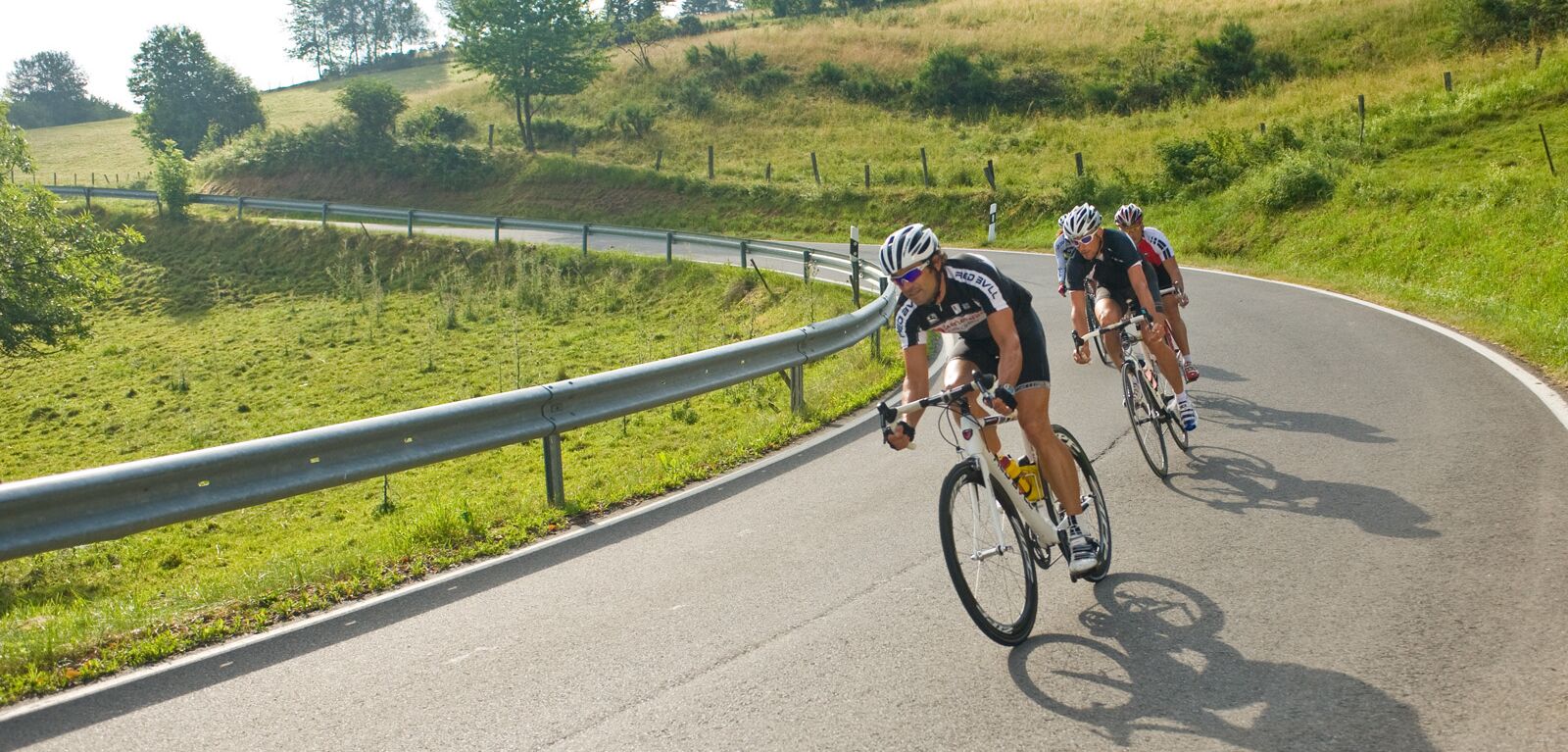 Rennradfahrer fahren rasant um eine Rechtskurve neben einem grün bewachsenen Hügel